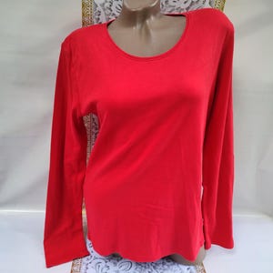 Könnte beinhalten: Ein langärmeliges, rotes Shirt mit Rundhalsausschnitt. Das Shirt wird auf einer Schaufensterpuppe präsentiert. Der Saum hat einen gewellten Rand. Der Stoff scheint einfarbig zu sein.