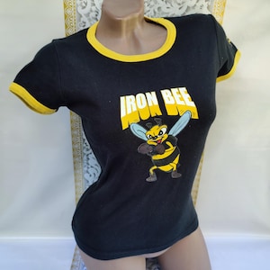 Può includere: Maglietta nera con bordino giallo e un'immagine di un'ape cartoon. Il testo "Iron Bee" è stampato sulla maglietta.