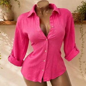 Può includere: Una camicia a maniche lunghe color rosa acceso con colletto classico, abbottonatura frontale con bottoni chiari e maniche arrotolate fino ai gomiti, esposta sul busto di un manichino.