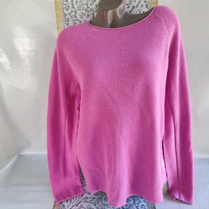 Puede incluir: Un suéter rosa brillante de punto acanalado con cuello barco y mangas largas. El suéter se muestra en un maniquí y tiene un corte holgado. La tela parece ser de peso medio, adecuada para climas más fríos. El estilo general es casual y cómodo.
