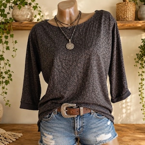 May include: A dark gray top with a subtle black leopard print pattern, featuring three-quarter length sleeves with folded cuffs and a wide neckline. The top is paired with blue denim shorts that have distressed details and a brown leather belt.