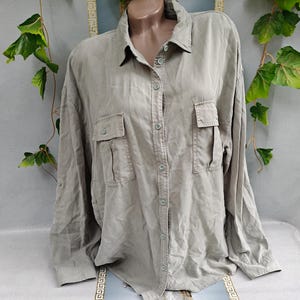 May include: A long-sleeved button-down shirt in a muted taupe color. The shirt features a classic collar, a full-length front button placket, and two chest pockets with buttoned flaps. The fabric has a soft, slightly wrinkled texture throughout.