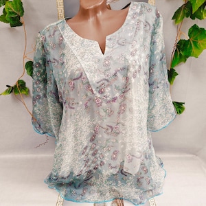 May include: A light blue tunic top with a V-neck and three-quarter sleeves. The sheer fabric features a floral pattern in shades of purple and white. The neckline and sleeve edges are trimmed with white lace and blue piping.