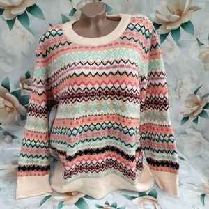 Puede incluir: Un suéter de color crema con un patrón geométrico colorido en tonos de rosa, azul, verde y negro. El suéter tiene un cuello redondo y mangas largas.