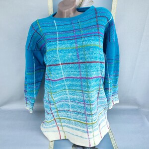 Puede incluir: Un suéter de manga larga en tonos azules, con un estampado de cuadros en rojo, morado y verde. El suéter tiene cuello redondo y un dobladillo inferior blanco. La prenda se exhibe en un maniquí.