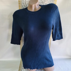Puede incluir: Top de punto acanalado azul marino con mangas cortas. El top tiene un cuello redondo y una silueta entallada. La tela parece ser de peso medio, adecuada para climas más frescos. Las mangas terminan justo por encima del codo.