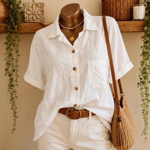 Puede incluir: Una camisa blanca de manga corta con botones, bolsillo en el pecho y botones marrones jaspeados, combinada con pantalones blancos, un cinturón de cuero trenzado marrón, un collar dorado de varias capas y un bolso de paja beige.