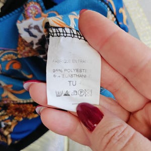 Puede incluir: Primer plano de una etiqueta con el texto "FABRIQUE EN FRANCE", "95% POLYESTER 5% ELASTHANE" y s&iacute;mbolos de lavado. La etiqueta se sostiene sobre una tela estampada azul y dorada.