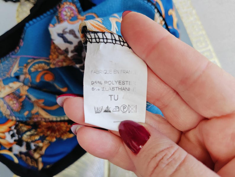 Puede incluir: Primer plano de una etiqueta con el texto "FABRIQUE EN FRANCE", "95% POLYESTER 5% ELASTHANE" y s&iacute;mbolos de lavado. La etiqueta se sostiene sobre una tela estampada azul y dorada.