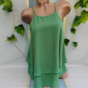 May include: A green, layered camisole top with thin spaghetti straps. The top is displayed on a mannequin, showcasing its flowing design. The fabric appears lightweight and suitable for warm weather. The top is a solid, vibrant green color.