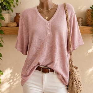 May include: A light pink short-sleeved shirt with a V-neckline and three buttons on the front placket. The fabric has a subtle textured pattern. The shirt is worn tucked into white pants with a brown leather belt and paired with a woven bag.