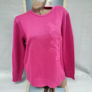 May include: A magenta crew neck sweater with long sleeves. The sweater has a textured floral design on the left side and a scalloped hem. The garment is displayed on a mannequin. The colour is a vibrant pink.