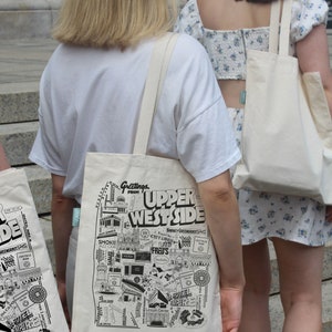 May include: Two people walking down a set of stairs, each carrying a white tote bag. The tote bag on the left has a black and white illustration of a city skyline with the text "Greetings from Upper West Side". The tote bag on the right is plain white.