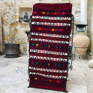 May include: A long, narrow, red, black, and white woven rug with geometric patterns and colorful accents. The rug has tassels on the ends and is hanging on a wall.