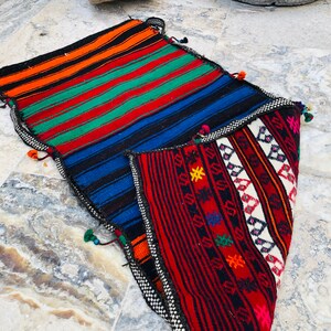 May include: A colorful woven rug with a geometric pattern in shades of red, blue, green, orange, and white. The rug is folded in half, revealing the intricate details of the design.