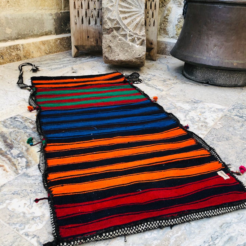 May include: A colorful striped rug with black, orange, red, blue, and green stripes. The rug has a fringe border and is made of woven fabric.