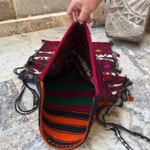 May include: A woven tapestry bag with a red, orange, green, and black striped interior. The bag is made of a thick, durable fabric and has a long, braided strap.