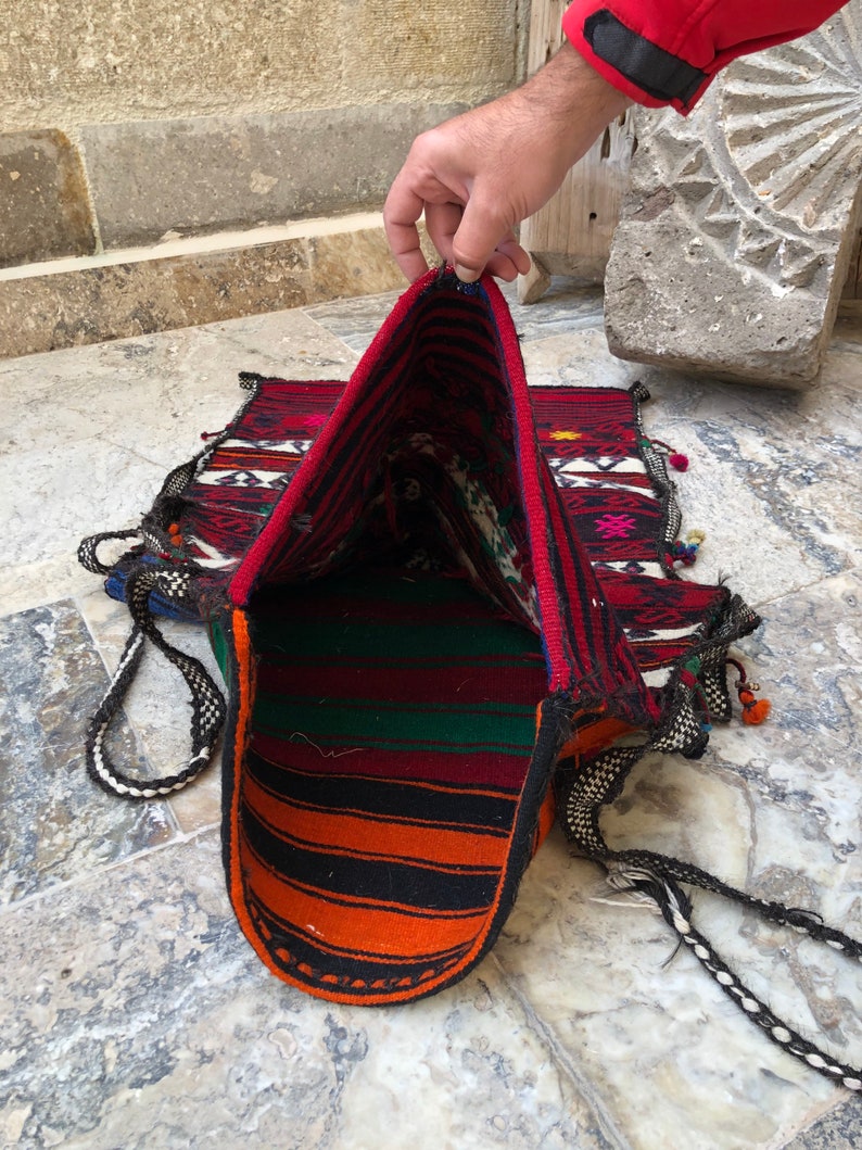 May include: A woven tapestry bag with a red, orange, green, and black striped interior. The bag is made of a thick, durable fabric and has a long, braided strap.