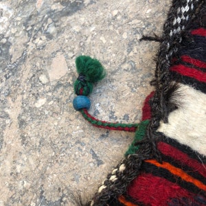 May include: Close-up of a colorful woven textile with a green tassel and a blue bead. The textile has a striped pattern in red, black, white, and orange.