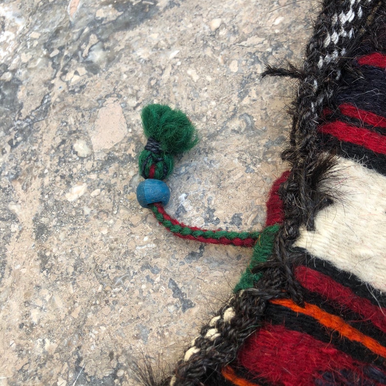 May include: Close-up of a colorful woven textile with a green tassel and a blue bead. The textile has a striped pattern in red, black, white, and orange.