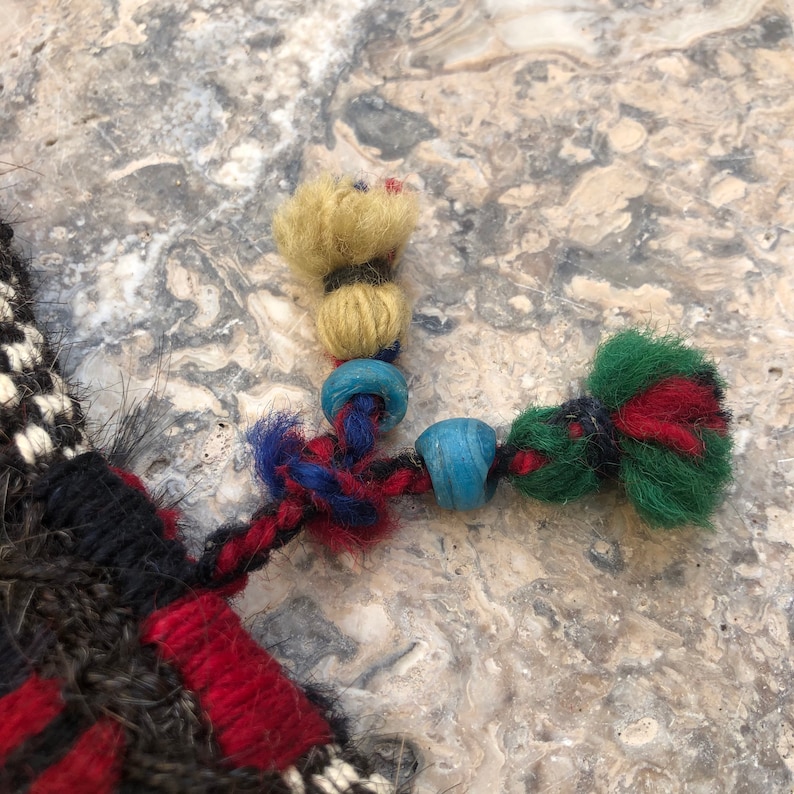 May include: A close-up of a braided cord with three colorful tassels. The tassels are made of yarn in shades of yellow, green, and red. The cord is decorated with blue and turquoise beads.