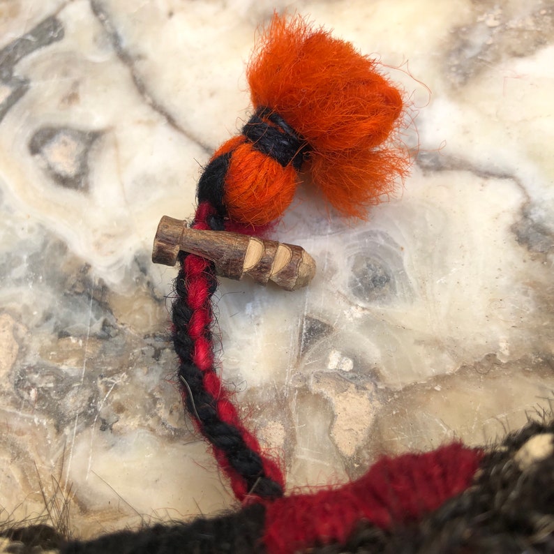 May include: A close-up of a red, black, and orange braided cord with a wooden toggle and a fluffy orange tassel.