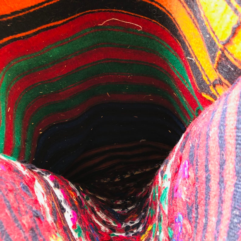 May include: A close-up of the inside of a woven rug. The rug is made of colorful stripes of red, green, orange, and black wool. The rug is folded in on itself, creating a tunnel-like shape.