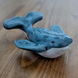 May include: A ceramic whale figurine with a blue and white glaze. The whale is sculpted in a realistic style with a detailed texture and a slightly open mouth.