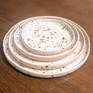 May include: Set of four white ceramic plates with brown speckles. The plates are stacked in a graduated size, with the largest plate on the bottom and the smallest plate on top.