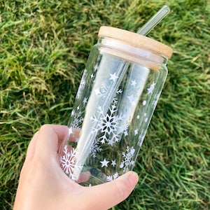 Puede incluir: Un vaso de vidrio transparente con tapa de madera y pajita de vidrio. El vaso está decorado con copos de nieve y estrellas blancas. El fondo es hierba verde.