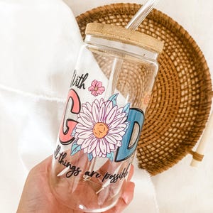 Puede incluir: Vaso de vidrio transparente con tapa y pajita de bambú. El vaso presenta las palabras "With God all things are possible" en un diseño colorido con una margarita y detalles florales.