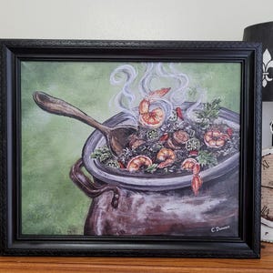 May include: A framed painting depicts a steaming pot of gumbo with shrimp, okra, and rice. A wooden spoon rests in the pot, and steam rises from the dish. The artwork is set against a green backdrop and framed in black.