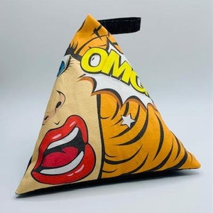 Pode incluir: Um calço de porta triangular com um design pop art que apresenta um rosto de mulher com uma expressão de surpresa e a palavra "OMG" em um balão de fala. O calço de porta é amarelo, laranja, vermelho, preto e branco.