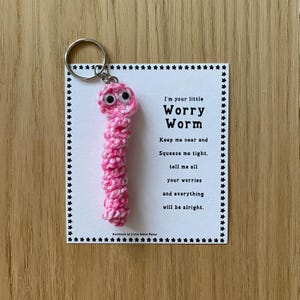 K&ouml;nnte beinhalten: Rosa geh&auml;kelter Sorgenwurm Schl&uuml;sselanh&auml;nger mit Glitzern und einer wei&szlig;en Karte mit der Aufschrift "Ich bin dein kleiner Sorgenwurm. Halte mich in deiner N&auml;he und dr&uuml;cke mich fest, erz&auml;hl mir all deine Sorgen und alles wird gut."