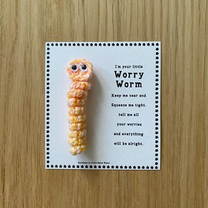 K&ouml;nnte beinhalten: Ein kleiner geh&auml;kelter orangefarbener und wei&szlig;er Sorgenwurm mit Glitz Augen. Der Wurm ist an einer wei&szlig;en Karte mit schwarzem Text befestigt, der lautet: "Ich bin dein kleiner Sorgenwurm. Halte mich in deiner N&auml;he und dr&uuml;cke mich fest, erz&auml;hl mir all deine Sorgen und alles wird gut."
