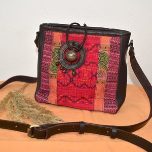 Könnte beinhalten: Eine braune Leder-Umhängetasche mit einer farbenfrohen bestickten Klappe. Die Klappe zeigt ein geometrisches Muster in Rot-, Rosa-, Grün- und Blautönen. Die Tasche hat eine Metallschnalle mit einem dekorativen Design.