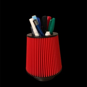 May include: A red cylindrical pencil holder with a ribbed design. The holder is filled with various pens and markers, including black, blue, green, and white.