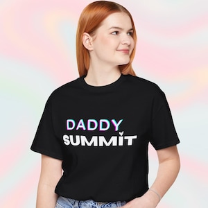 May include: Black t-shirt with the text "DADDY SUMMIT" in white, with a blue and pink outline. The model is wearing blue jeans. The background is a colorful, blurred pattern.