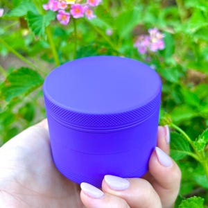 Puede incluir: Un molinillo de metal morado con una tapa texturizada. El molinillo se est&aacute; sosteniendo en una mano.