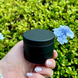 May include: A black metal grinder with four sections. The grinder is held in a hand with white nail polish.