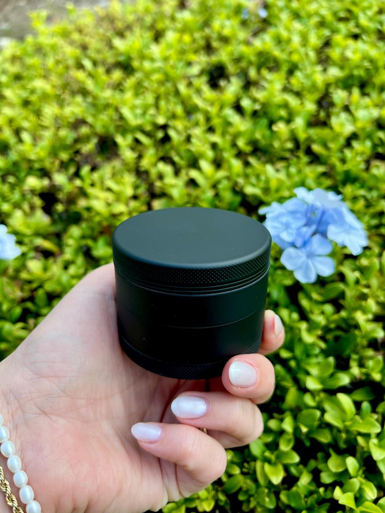 May include: A black metal grinder with four sections. The grinder is held in a hand with white nail polish.