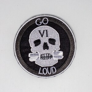 May include: A black and white embroidered patch with a skull and crossbones design. The skull has the Roman numeral VI on its forehead and the words "GO LOUD" around it.