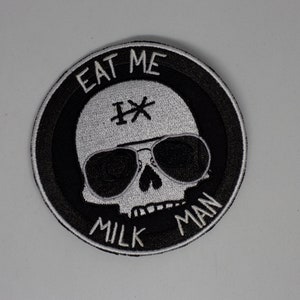 May include: A black and white embroidered patch with a skull wearing sunglasses and a hat that says "Eat Me Milk Man".