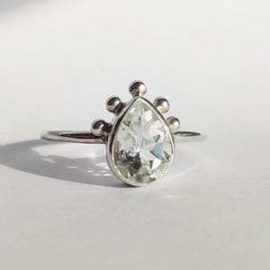 Könnte beinhalten: Ein silberner Ring mit einem birnenförmigen, klaren Edelstein. Der Edelstein ist von einer silbernen Fassung mit kleinen, runden Akzenten umgeben. Der Ring ist vor einem weißen Hintergrund mit Schatten links platziert.