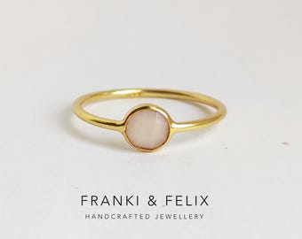 Könnte beinhalten: Ein goldfarbener Ring mit einem runden, hellrosa Edelstein in der Mitte. Der Ring hat ein zartes Band und wird auf einer weißen Oberfläche präsentiert. Unter dem Ring steht der Text "FRANKI & FELIX HANDCRAFTED JEWELLERY".