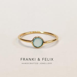 Può includere: Un delicato anello d'oro con una gemma rotonda azzurra al centro. L'anello ha una fascia sottile e un design semplice ed elegante. Il testo "FRANKI & FELIX HANDCRAFTED JEWELLERY" è stampato sotto.