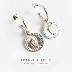 May include: Silver hoop earrings with a coin charm featuring a profile of a person's face. The coin has a raised rim and intricate details.