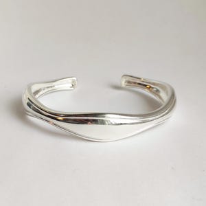 May include: A silver cuff bracelet with a smooth, flowing design. The open-ended bracelet has a curved shape with a wider centre section and tapers towards the ends. The polished metal reflects light, creating highlights and shadows.