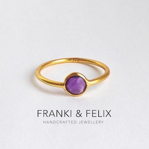 May include: A gold ring featuring a round, purple gemstone. The ring has a thin gold band. The text "FRANKI & FELIX HANDCRAFTED JEWELLERY" is printed below the ring. The ring is a piece of handcrafted jewellery.