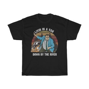 May include: A black t-shirt featuring a retro-style graphic of a man in a plaid shirt standing next to a yellow van. The graphic includes the text "Livin in a Van Down by the River".